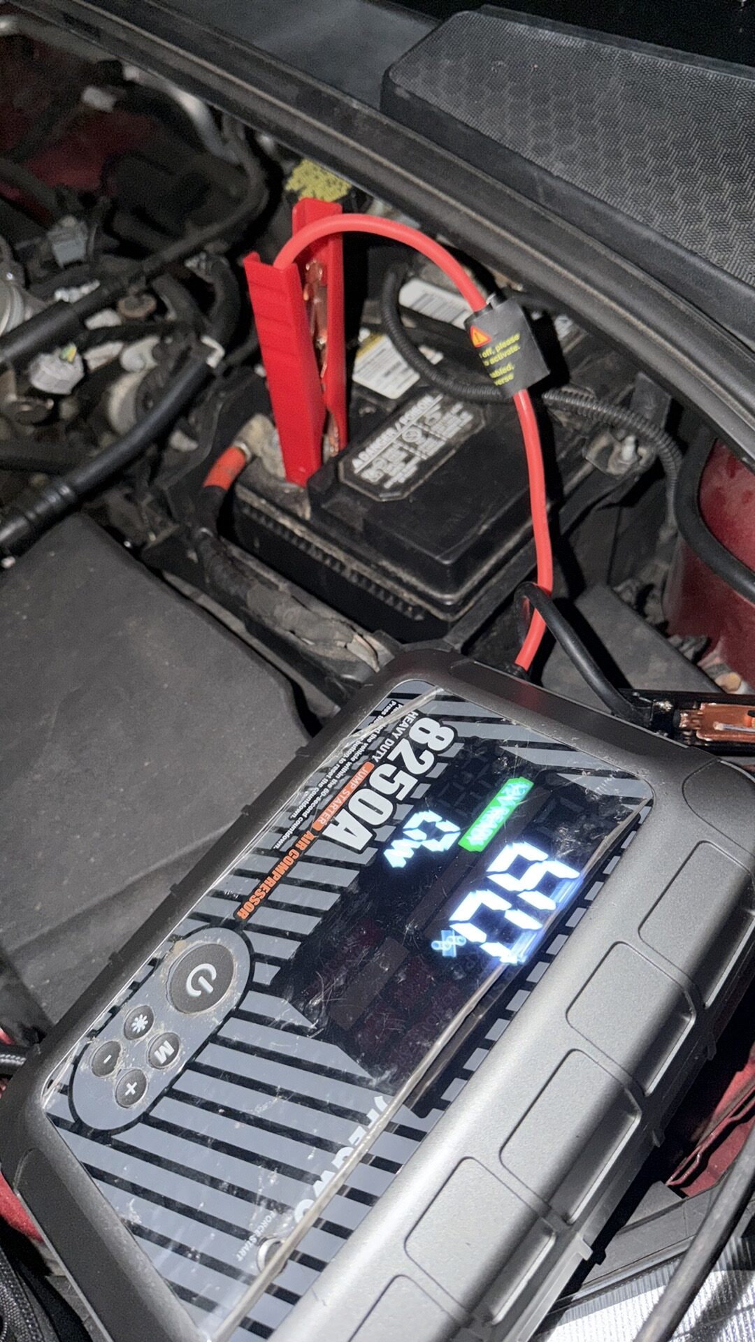 Close-up of an 8250A-labelled portable jump starter with its display illuminated beside a vehicle battery