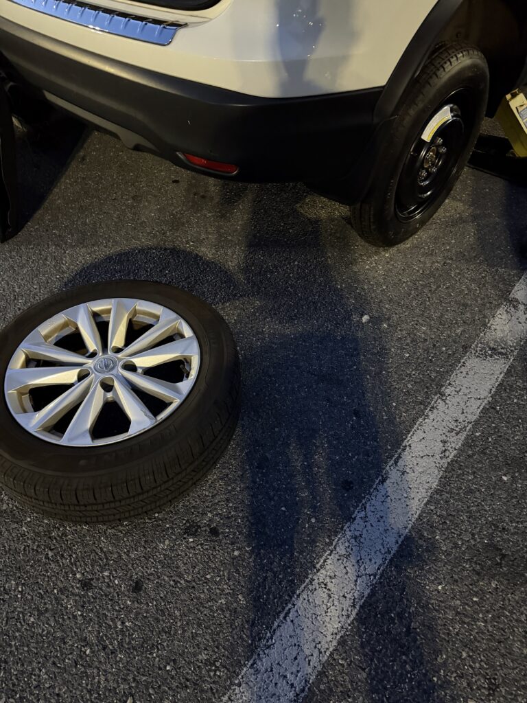 Temporary spare fitted to a white Nissan Rogue with the removed road wheel beside the vehicle on Security Boulevard