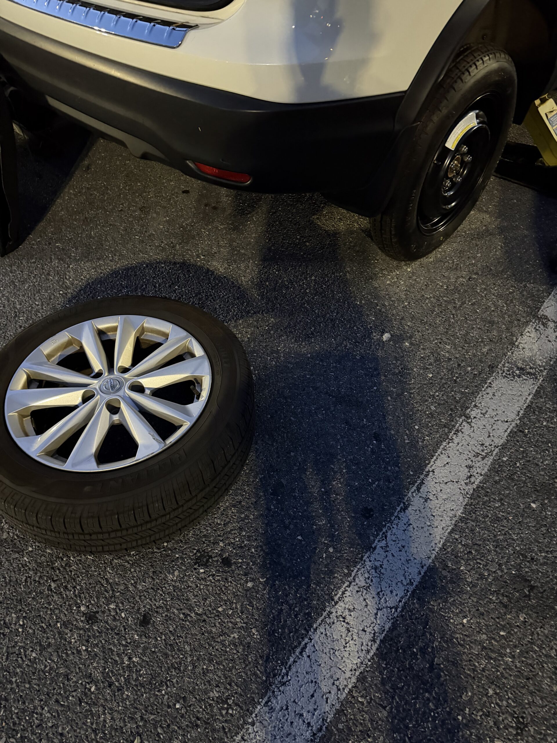 Temporary spare fitted to a white Nissan Rogue with the removed road wheel beside the vehicle on Security Boulevard
